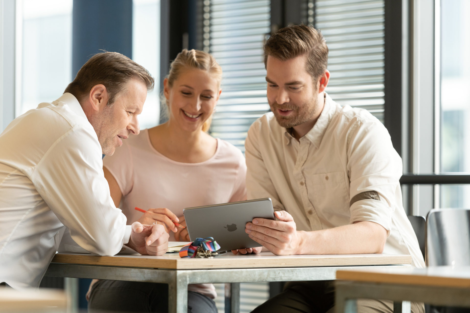 Zwei Kursteilnehmende und ein Dozent schauen auf ein Tablet. Alle sitzen um ein Pult herum.