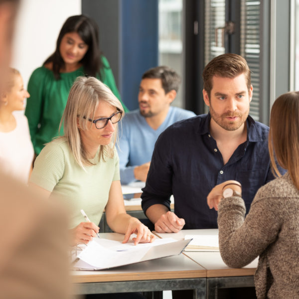 Mehrere Kursteilnehmer*innen sitzen um ein Pult und diskutieren miteinander. Auf dem Pult sind Ordner und verschiedene Blätter platziert. Im Hintergrund sind weiter Kursteilnehmer*innen in der Unschärfe.