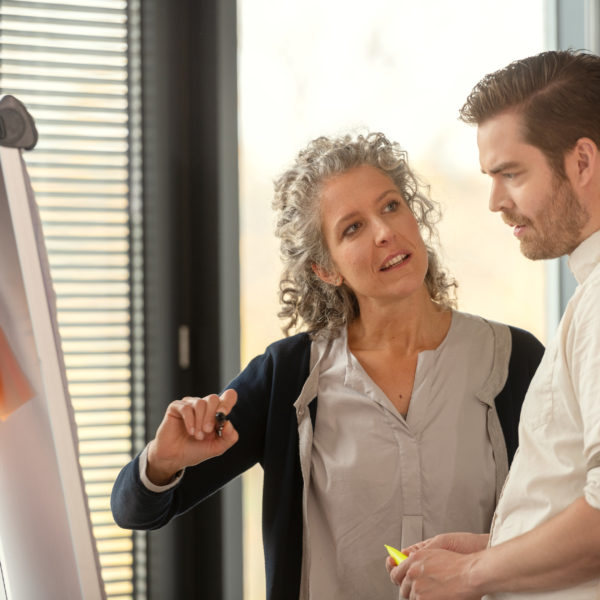 Eine Kursteilnehmerin und ein Kursteilnehmer vor einem Flip Chart miteinander am diskutieren. Im Hintergrund sind die Fenster des Klassenraums sichtbar.