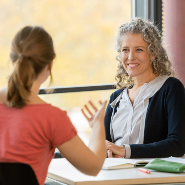 An einem Pult sitzen eine Dozentin und Kursteilnehmerin und diskutieren mtieinander. Im Hintergrund ist eine farbige Säule und die Fenster des Klassenraums.