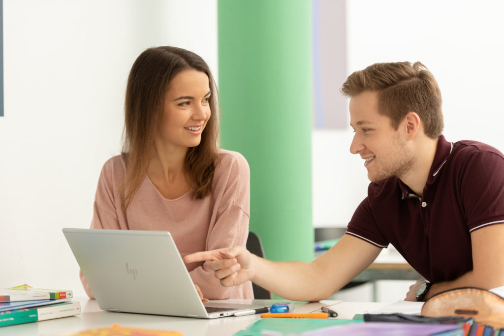 Zwei Lernende an einem Pult vor einem Laptop sitzend. Auf dem Pult sind Bücher, Schreibzeug , farbige Mäppli verteilt. Im Hintergrund hat es eine grüne Säule sowie ein blaues Bild