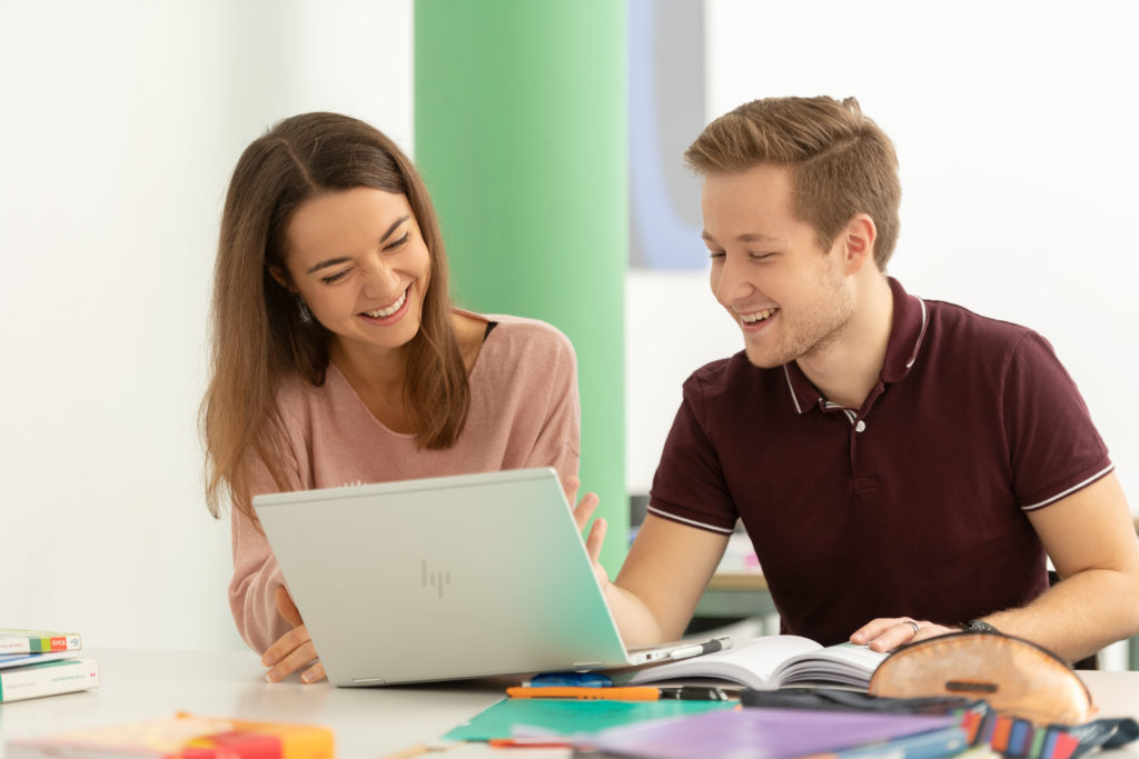 Zwei Lernende an einem Pult vor einem Laptop sitzend. Auf dem Pult sind Bücher, Schreibzeug , farbige Mäppli verteilt. Im Hintergrund hat es eine grüne Säule sowie ein blaues Bild