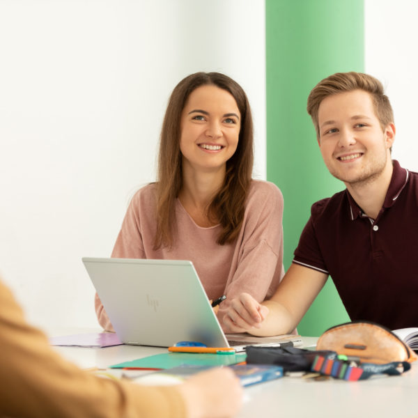 Drei Lernende am Pult sitzend schauen nach vorne zur Lehrperson. Auf dem Pult sind Bücher sowie Laptop verteilt. Im Hintergrund ist eine grüne Säule sowie ein blaues Bild.