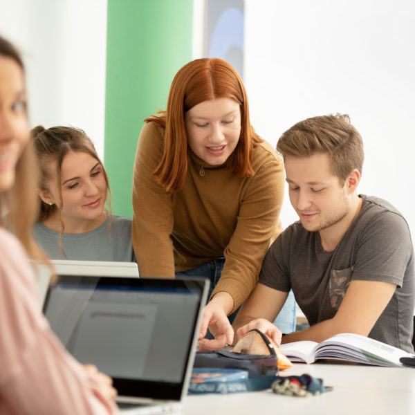 Vier Lernende um ein Pult sitzend schauen miteinander in Bücher und Laptop. Im Hintergrund eine gründe Säule und Bilder.