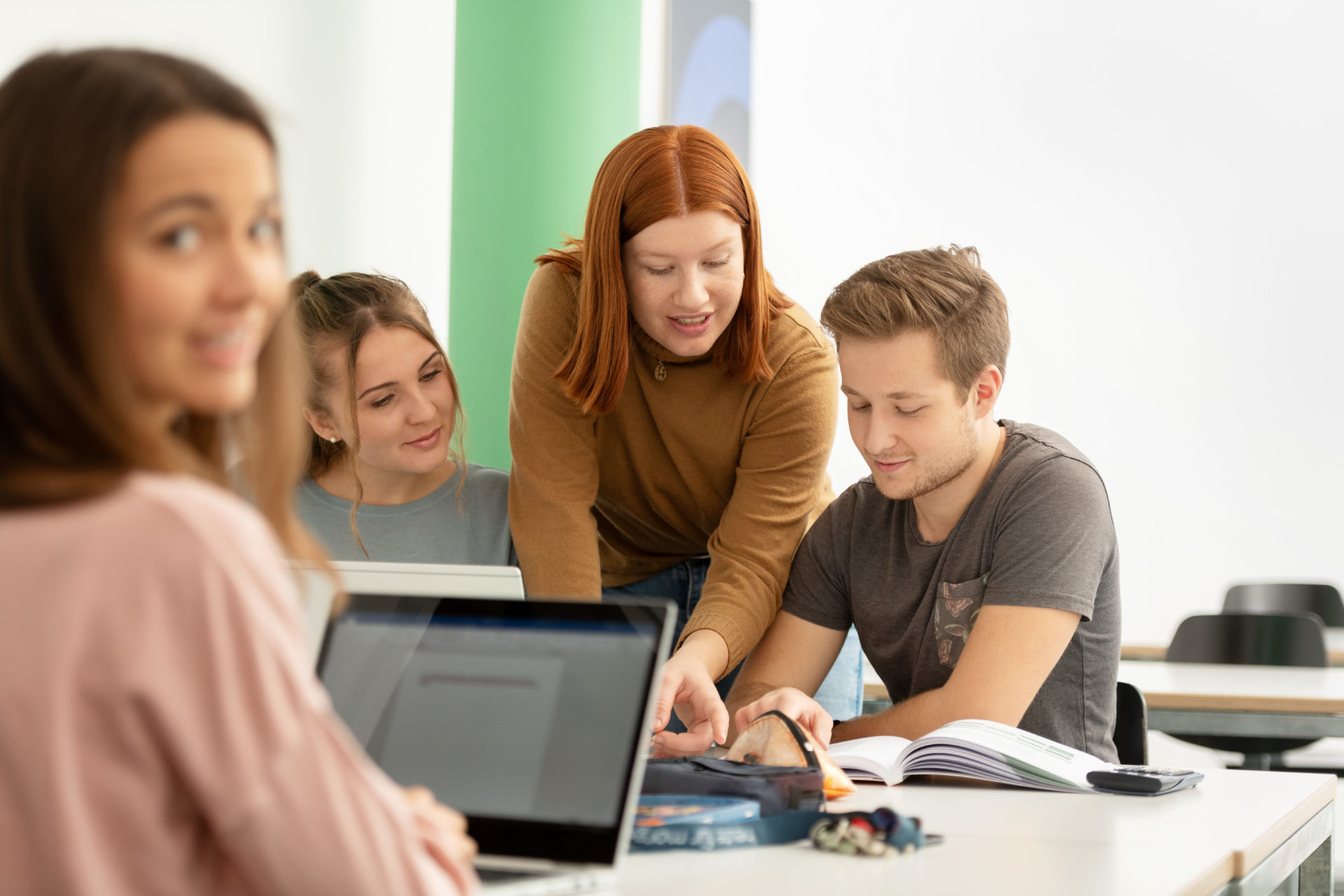 Vier Lernende um ein Pult sitzend schauen miteinander in Bücher und Laptop. Im Hintergrund eine gründe Säule und Bilder.
