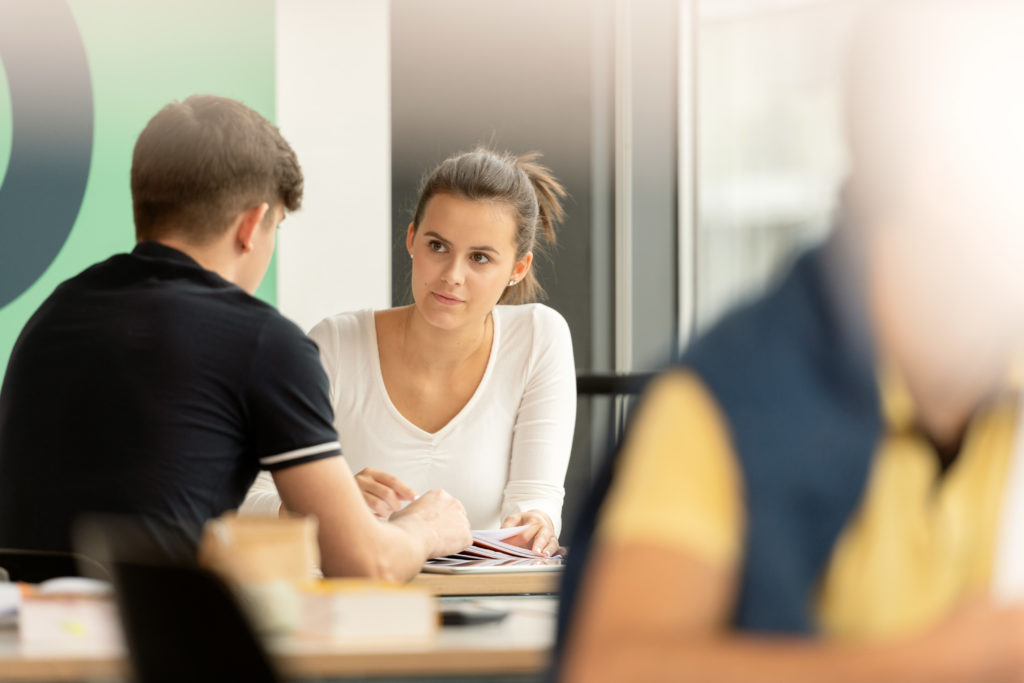 Im Hintergrund zwei Lernende mit offenen Bücher an einem Pult. Im Vorderrund in der Unschärfe ein Lernender am Lesen.