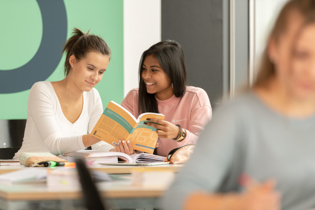 Im HIntergrund zwei Lernende die zusammen in ein Lernbuch schauen und im Vordergrund eine Lernende am schreiben.
