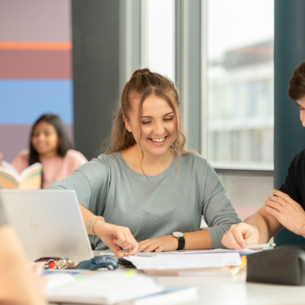 Lernende um ein Pult herum platziert, die auf Aufgaben-Blätter schauen.
