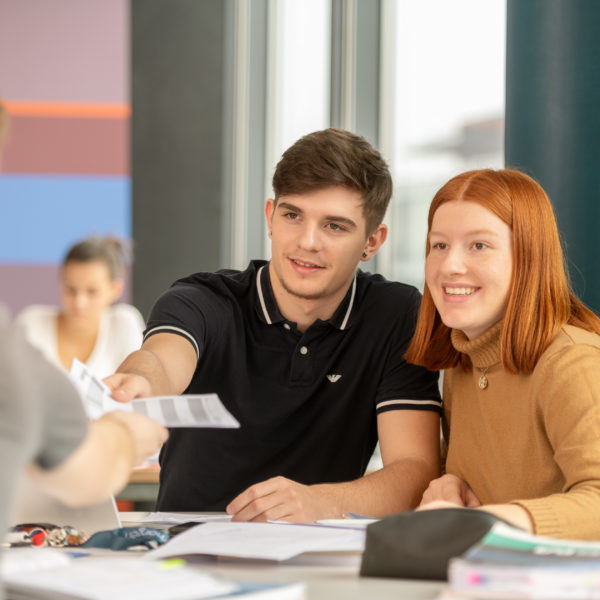 Lernende um ein Pult herum platziert, die untereinander Bläter austauschen.