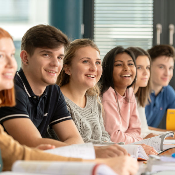 Sieben Lernende im Klassnzimmer, am Pult sitzen und nach vorne zum Lehrer schauen..