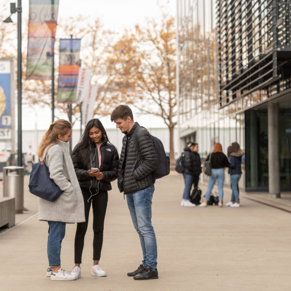 In der Mitte sind mehrere Lerndende die vor dem bwd Schulgebäude das sich am rechten Rand befindet, stehen.