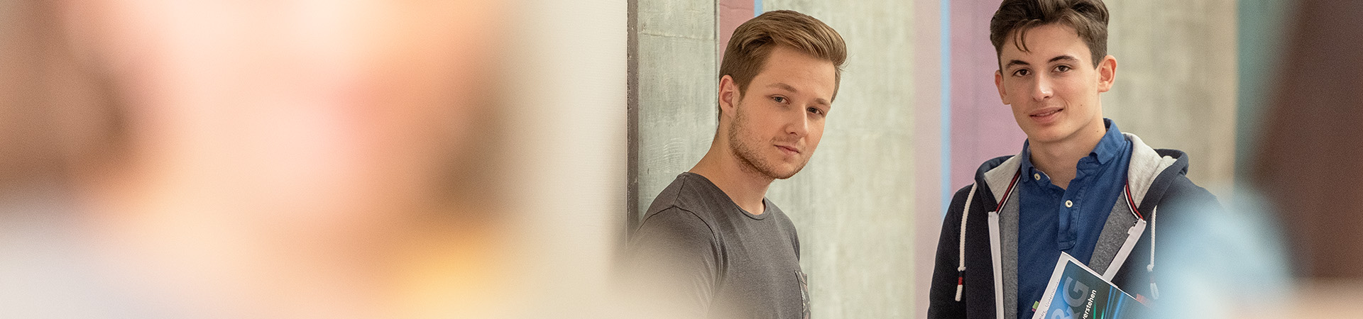 Zwei Lernende im Vordergrund in Unschärfe am Lernen und zwei Lernende im Hintergrund vor einem Laptop am diskutieren. Alle stehen im Gang vor den Klassenzimmern.