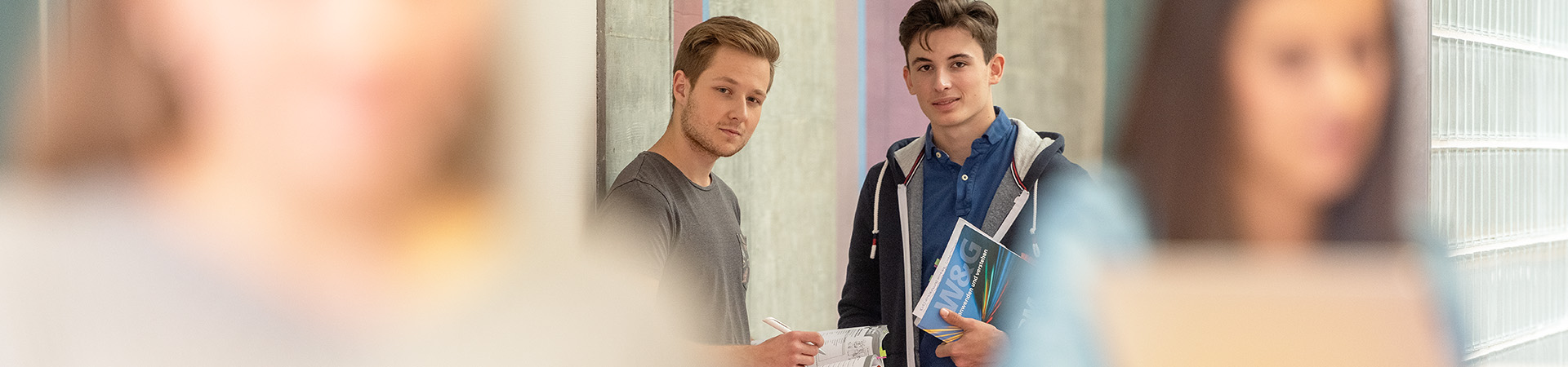 Zwei Lernende im Vordergrund in Unschärfe am Lernen und zwei Lernende im Hintergrund vor einem Laptop am diskutieren. Alle stehen im Gang vor den Klassenzimmern.