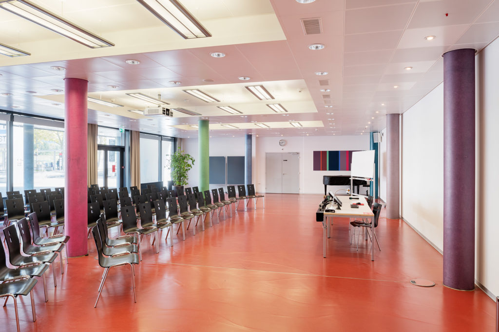 Saal 016. Link mit schwarzen Stühlen in Konzertbestuhlung. Links Tisch mit Laptop. An der linken Wand sind zwei farbige Säulen.