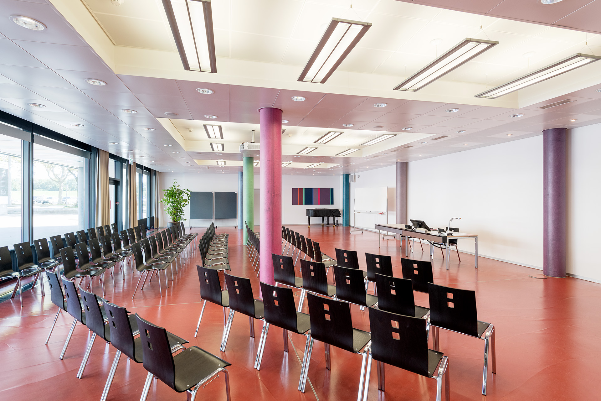 Saal 016. Grosser Raum mit Konzertbestuhlung mit schwarzen Stühlen. Rechts ist eine Fenserfront. Im Hintergrund Notizboards und ein farbiges Bild sowie ein Flügel. Rechts vor der weissen Wand 3 farbige Säulen und ein Tisch mit Laptop.