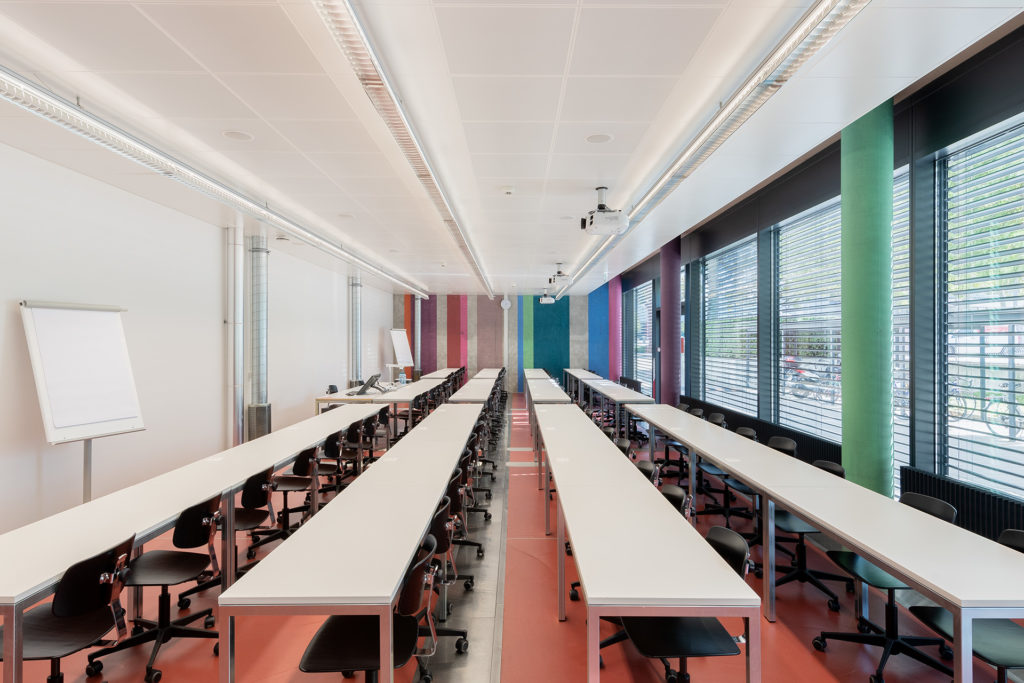 Kursraum 018 In der Mitte sind 4 lange Tischreihen in weiss mit schwarzen Stühlen davor. links eine weisse Wand mit zwei Flip-Charts. Im Hintergrund eine Betonwand mit farbigen Streifen. Rechts ist eine Fensterfront.