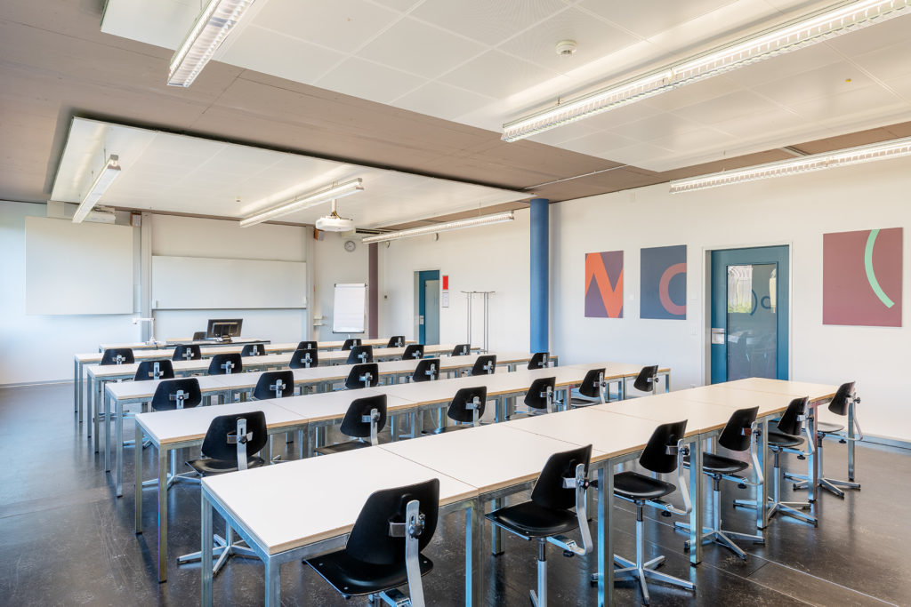 Klassenzimmer mit 24 Arbeitsplätzen. In der MItte mehrere Reihen weisse Tische hintereinander und davor schwarze Stühle. Hintergrund ist ein Whiteboard. Rechts zwei Ein-/Ausgänge sowie farbige Bilder.