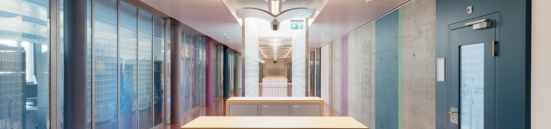 Schulzimmergang. Glaswand mit verschiedenen farbigen Säulen davor. Rechter Rand Klassenzimmertür. In der Mitte, Gepäckaufbewahrungsboxen sowie Treppenhaus.