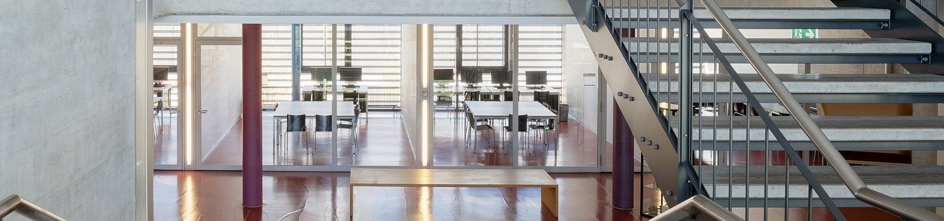 Zwei Gruppenräume für Lernende/Kursteilnehmer. Vom Treppenhaus aus zwei Gruppenräume ersichtlich, die je ein weisser Tisch mit Stühlen und je ein weisser Tisch mit 2 PCs haben. Vor den Gruppenräumen zwei farbige Säulen, dazwischen eine Holzbank.