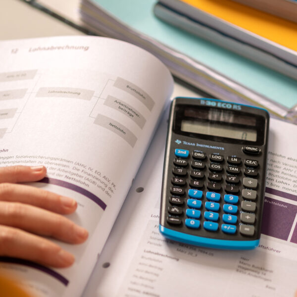 Rechts unten eine Hand die die Blätter eines Buches dass mittig liegt festhält. Auf dem Buch ist ein Rechner. Am oberen Rand sind Bücher und Hefte aufeinandergestapelt.