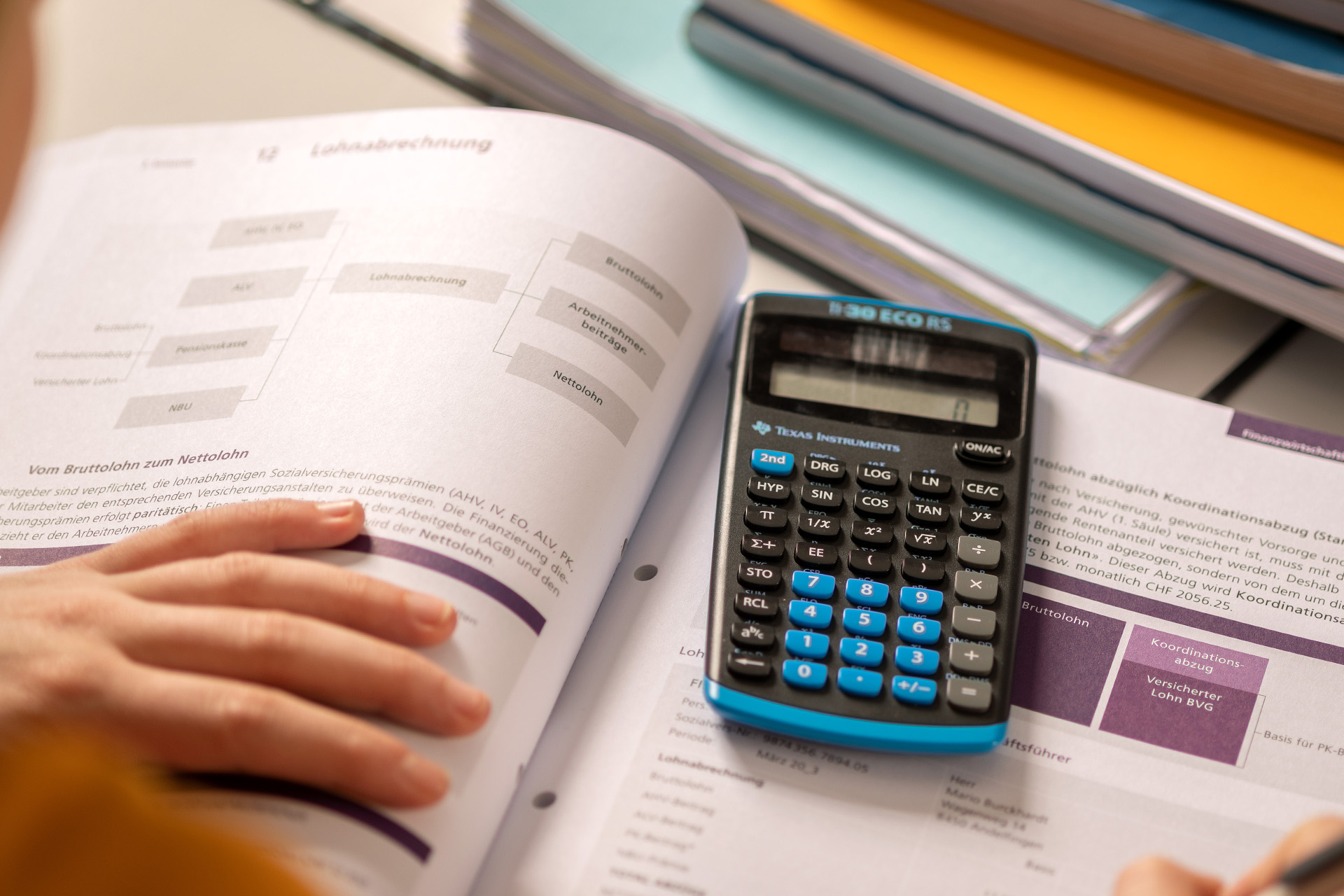 Rechts unten eine Hand die die Blätter eines Buches dass mittig liegt festhält. Auf dem Buch ist ein Rechner. Am oberen Rand sind Bücher und Hefte aufeinandergestapelt.
