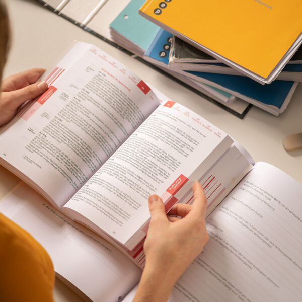 Links angeschnitten eine Lernende die in einem Schulbuch blättert. Oben rechts Schulbücher und Hefter aufeinander gestapelt.