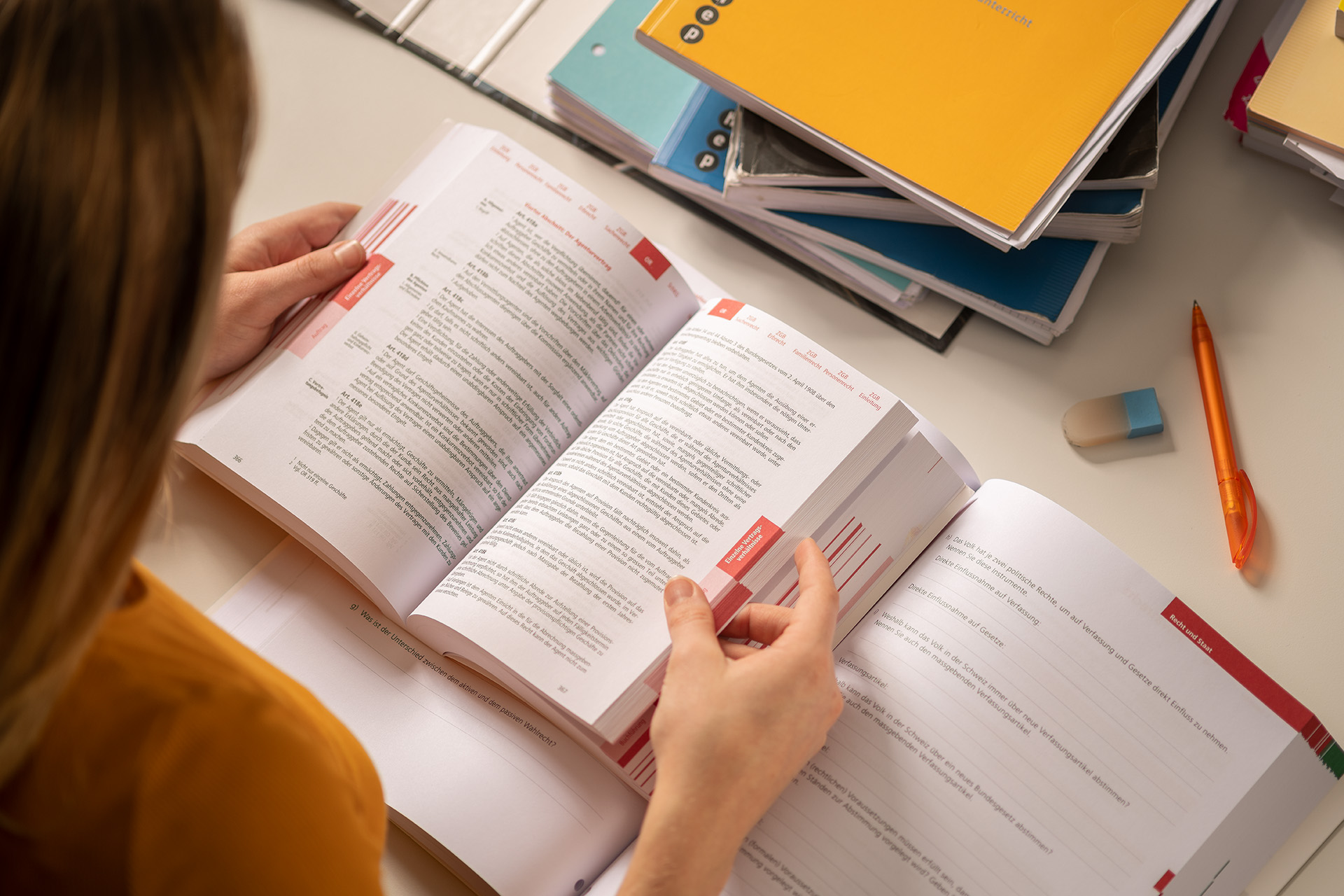 Links angeschnitten eine Lernende die in einem Schulbuch blättert. Oben rechts Schulbücher und Hefter aufeinander gestapelt.