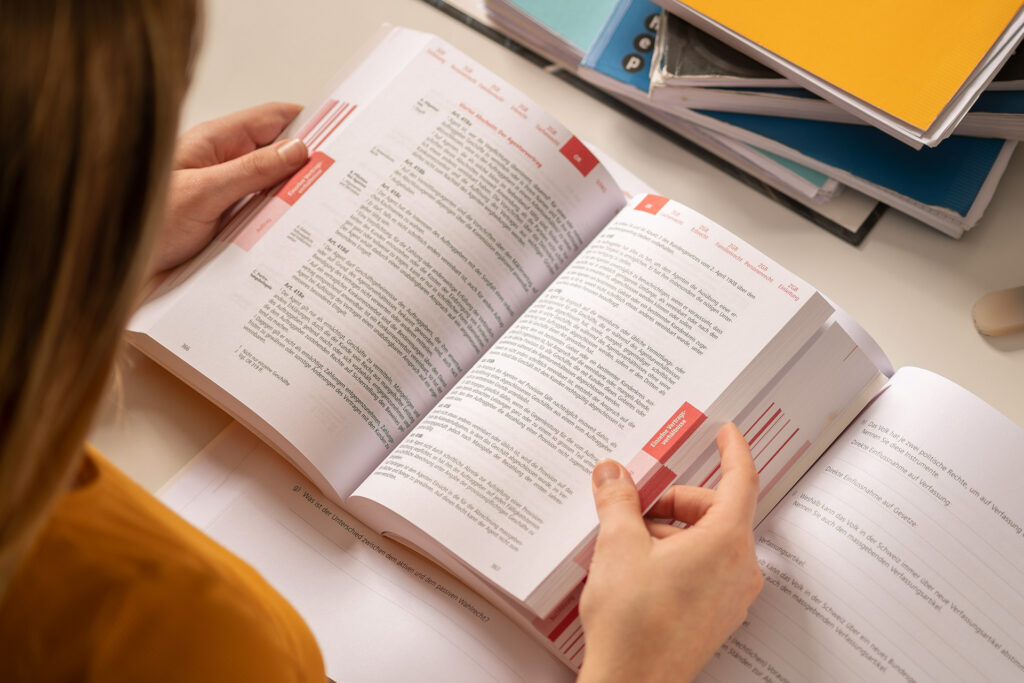 Links angeschnitten eine Lernende die in einem Schulbuch blättert. Oben rechts Schulbücher und Hefter aufeinander gestapelt.