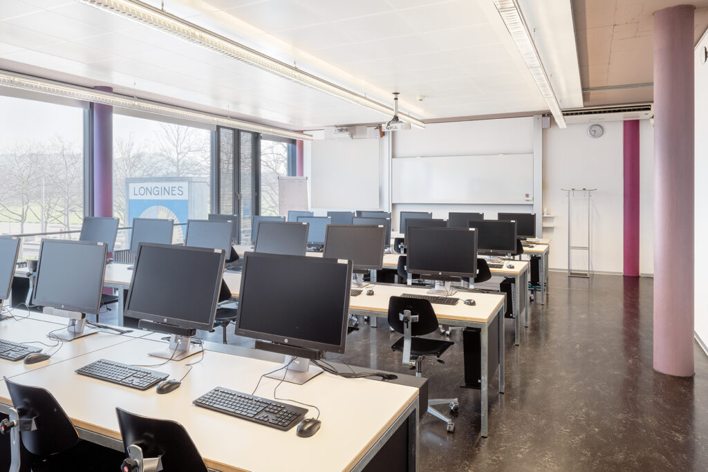 Informatikzimmer: Vor einer Fensterfront mit Sicht auf YB-Uhr sind 4 Reihen Tische mit mehreren Bildschirme, Tastatur und Stühlen ersichtlich. Rechts hat es eine pink und violettefarbige Säulen. Im Hintergrund ist eine weisse Wandtafel.