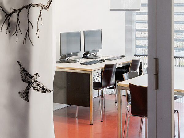 Gruppenraum für Lernende/Studenten. Ein Tisch mit 6 Stühlen in der Mitte des Raums, am linken Rand 1 Tisch mit 2 PC's. Hintergrund eine Fensterwand.