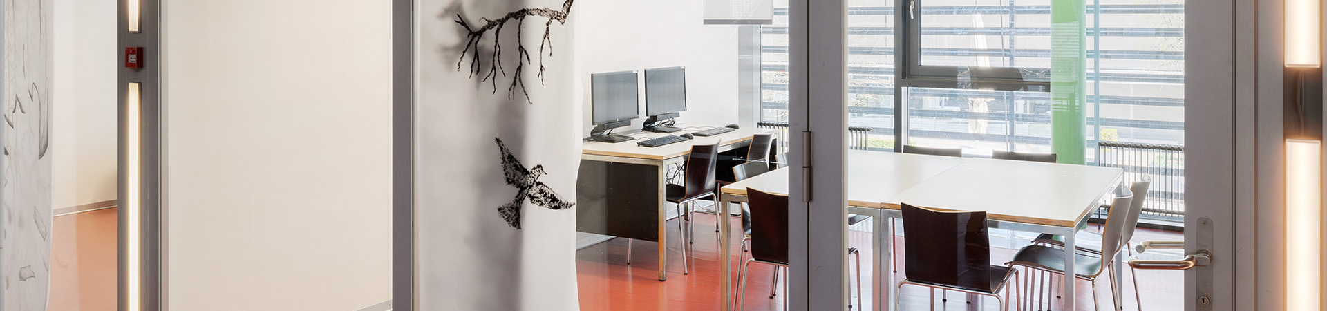 Gruppenraum für Lernende/Studenten. Ein Tisch mit 6 Stühlen in der Mitte des Raums, am linken Rand 1 Tisch mit 2 PC's. Hintergrund eine Fensterwand.