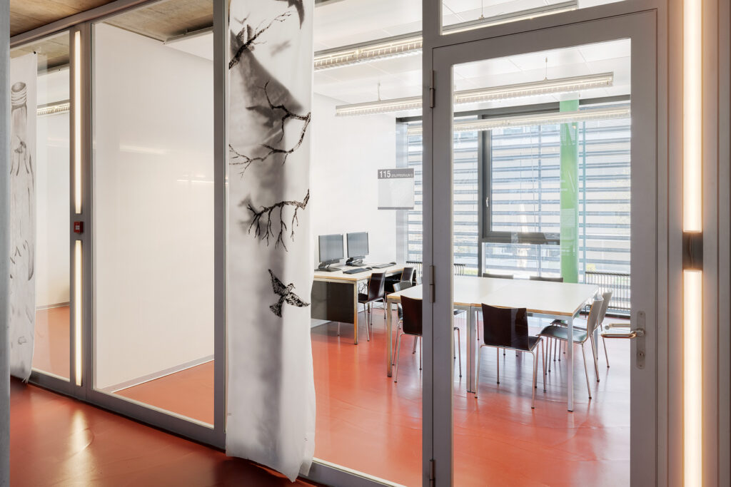 Gruppenraum für die Lernenden/Kursteilnehmer. Ein Tisch mit 8 stühlen und 1 Tisch mit 2 PC. Roter Boden. Front und Rückseite sind aus Glas. .