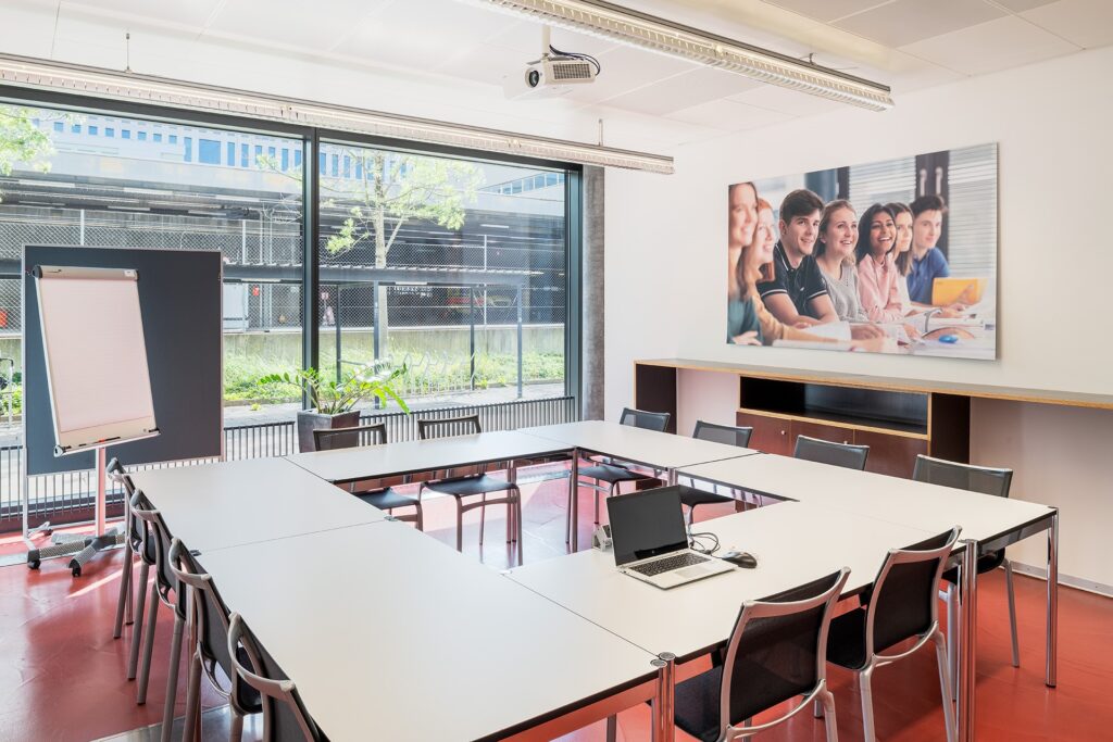 Sitzungszimmer. Tische im Quadrat platziert. 12 bis 16 Sitzplätze. Bild mit 6 Stundente. Rückwand ist eine Fensterwand.