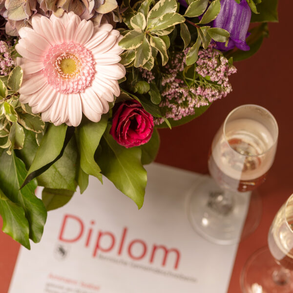 Linker oberer Rand Blumenstrauss mit rosa Blumen. Unter Rand Diplom mit zwei gefüllten Champagnergläser.